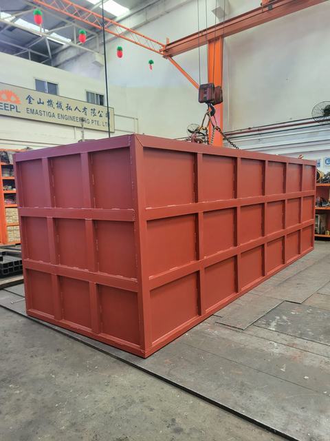 Red-painted steel box structure lifted by an overhead crane in the Emastiga workshop